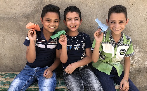 three boys sit on a bench, smiling and holding paper airplanes